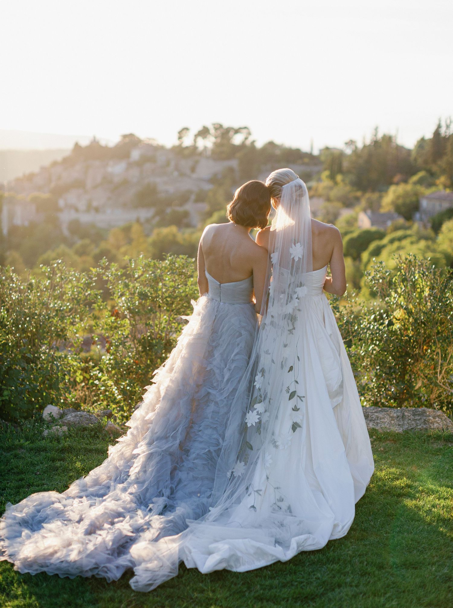 A romantic wedding couple sits together in a sunlit field wearing white gowns as golden light filters through the trees.