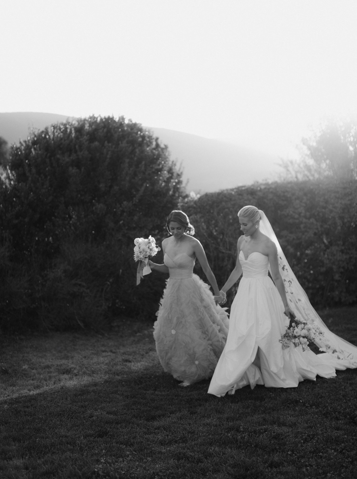 Two brides in flowing white dresses sit together on a grassy hillside at sunset in a romantic black and white photo.