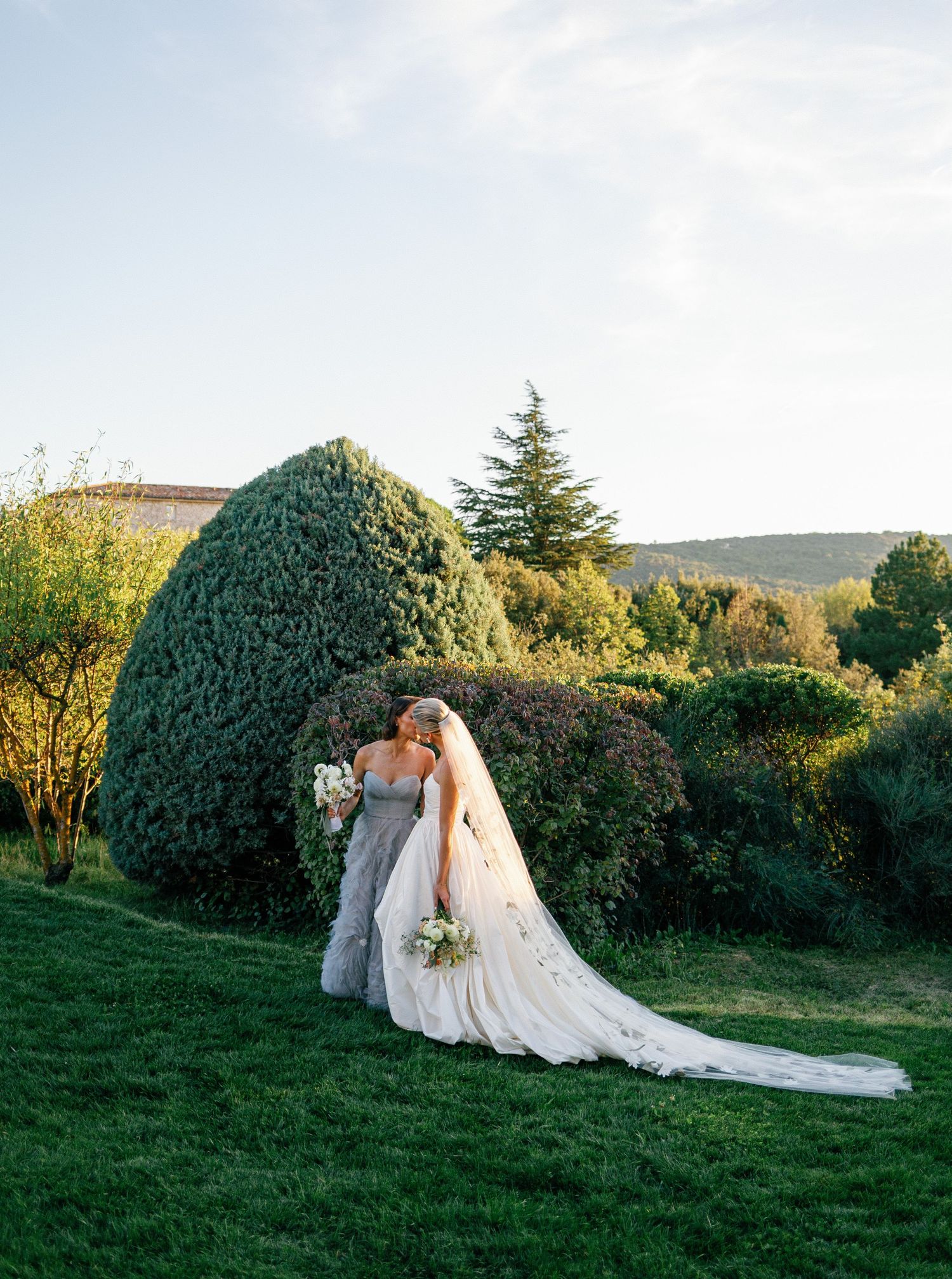 A couple in wedding attire embrace on a manicured lawn with ornamental shrubs and mountains in the background.