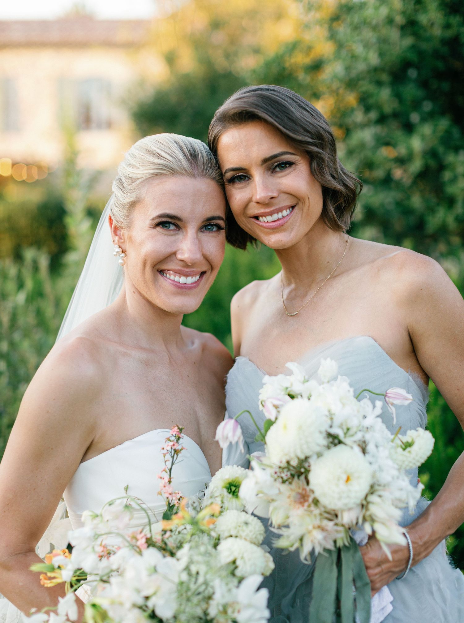 Two people in elegant wedding attire hold white floral bouquets while sharing a joyful moment outdoors at sunset.