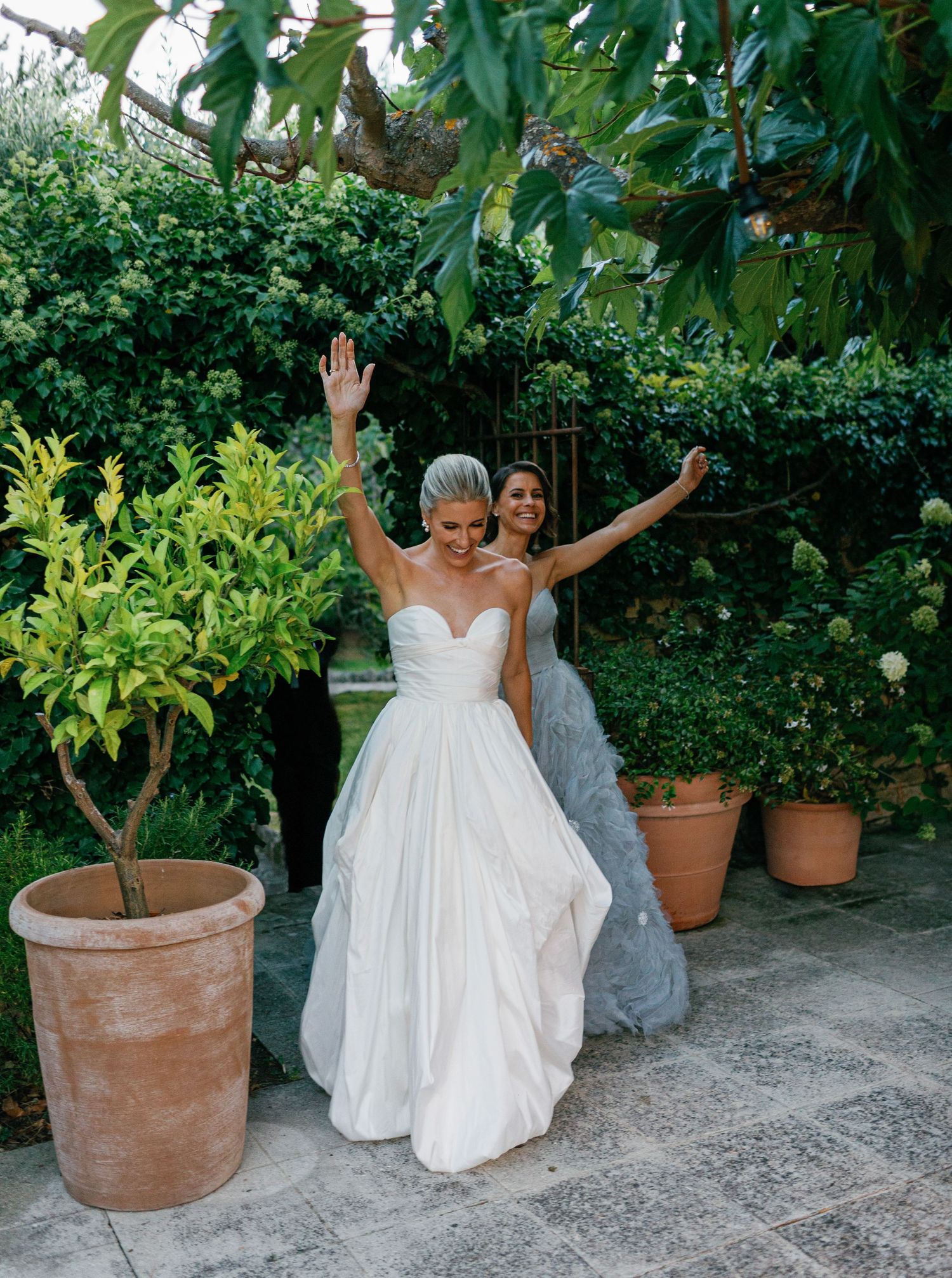 A joyful couple in wedding attire poses playfully in a lush garden setting with terracotta planters.