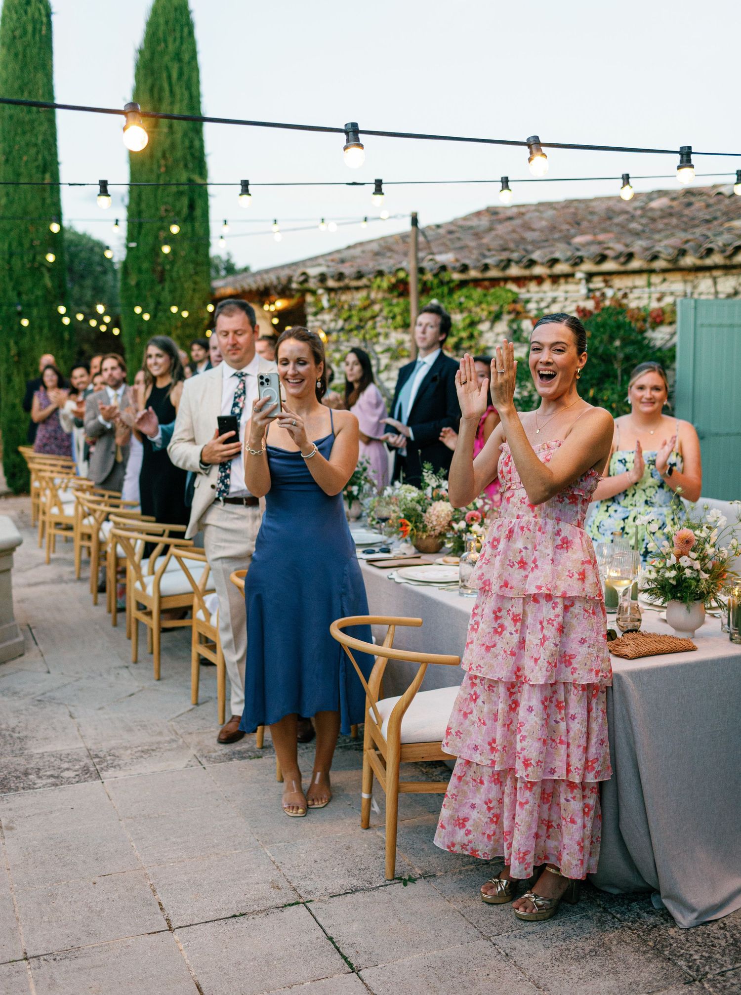Outdoor wedding reception guests seated at long tables under string lights with cypress trees in the background.