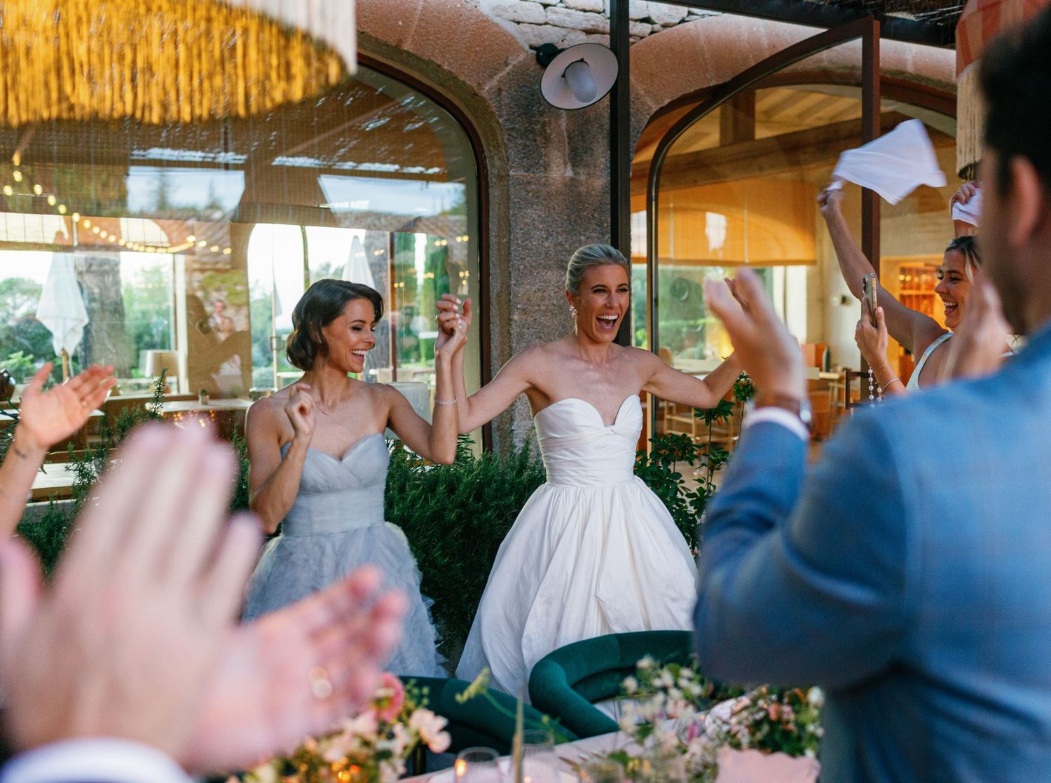 Wedding guests raise their arms in celebration during an outdoor reception with string lights overhead.
