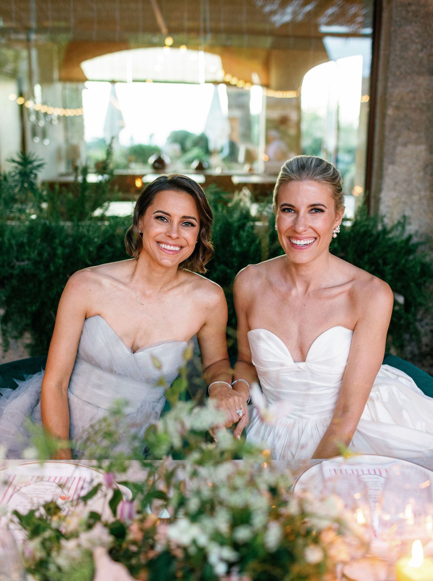 Two bridesmaids in strapless gowns share a candid moment at an elegant table setting with floral centerpieces.
