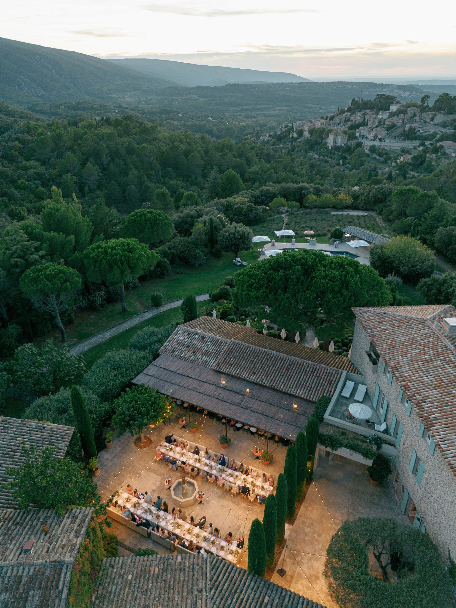 Aerial drone view of an illuminated rustic courtyard venue in a scenic mountain landscape at dusk.