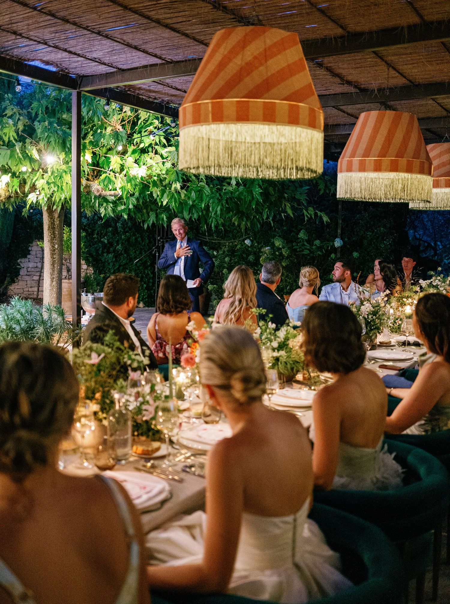 Wedding guests seated at a long table under a rustic pergola during an elegant outdoor dinner reception.