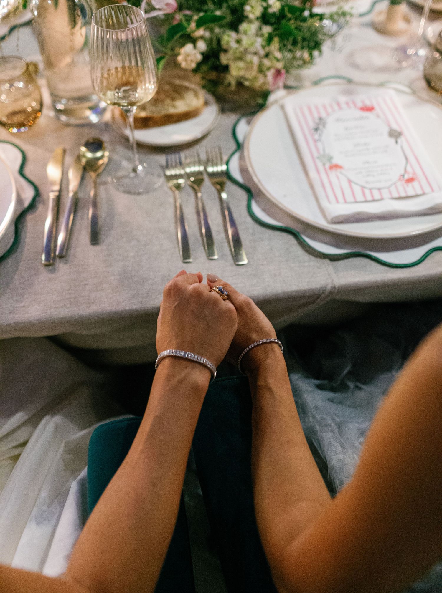Hands wearing delicate bracelets rest on an elegant table setting with fine china and glassware at a formal dinner.