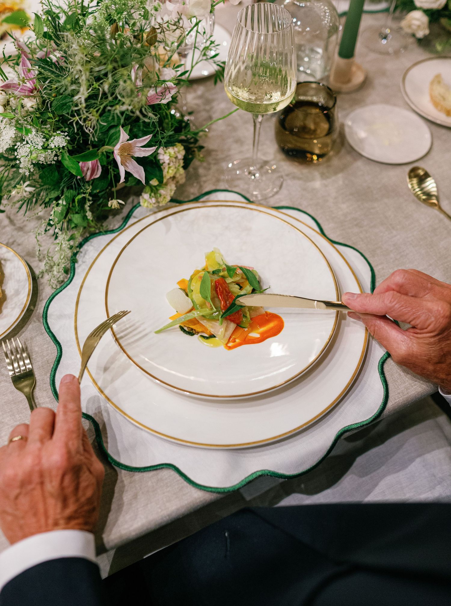 Elegant table setting with white and gold-rimmed plates, fresh floral centerpiece, and hands serving a gourmet dish.