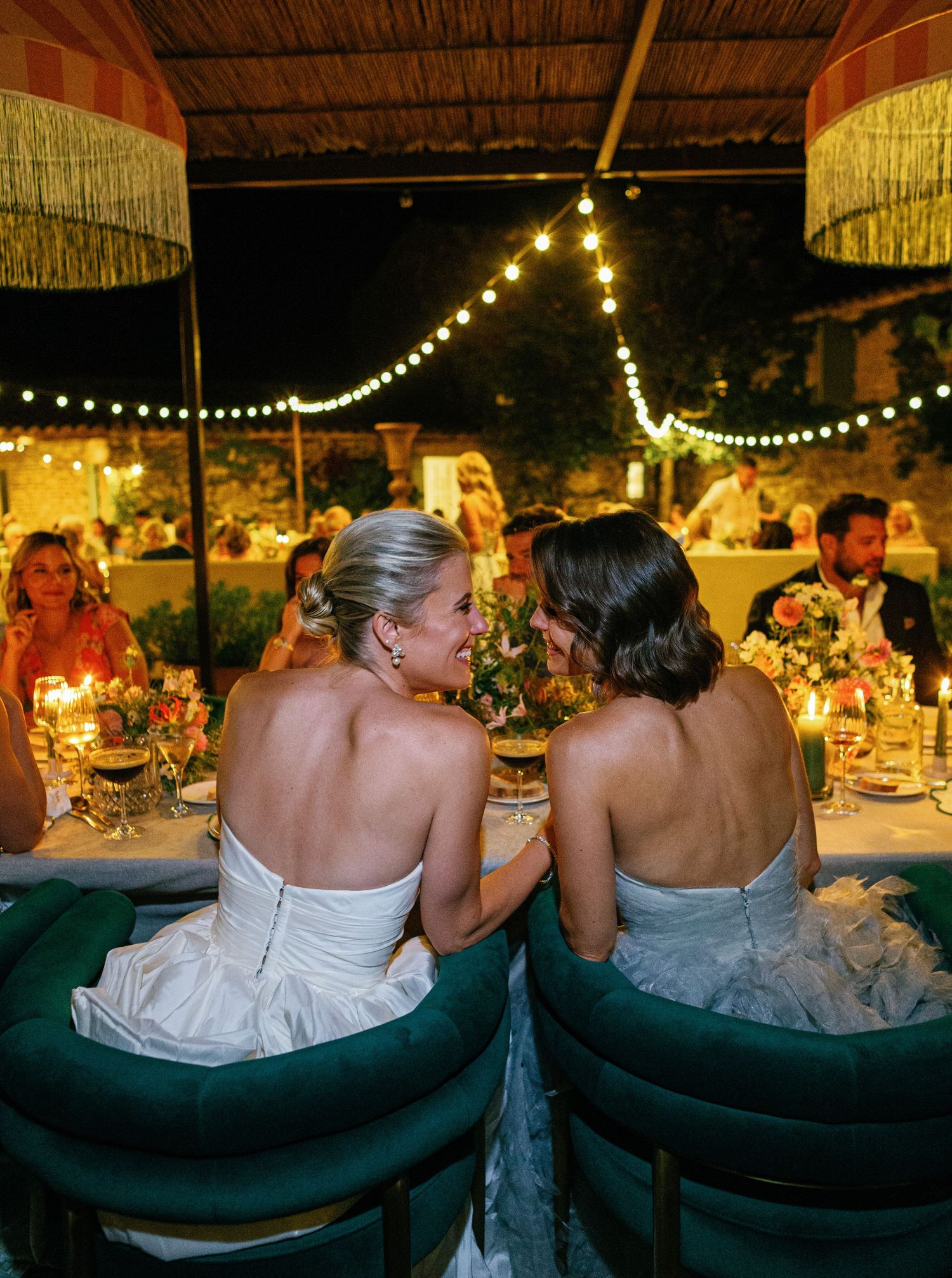 Wedding guests seated at a rustic outdoor reception under twinkling string lights and thatched roof.