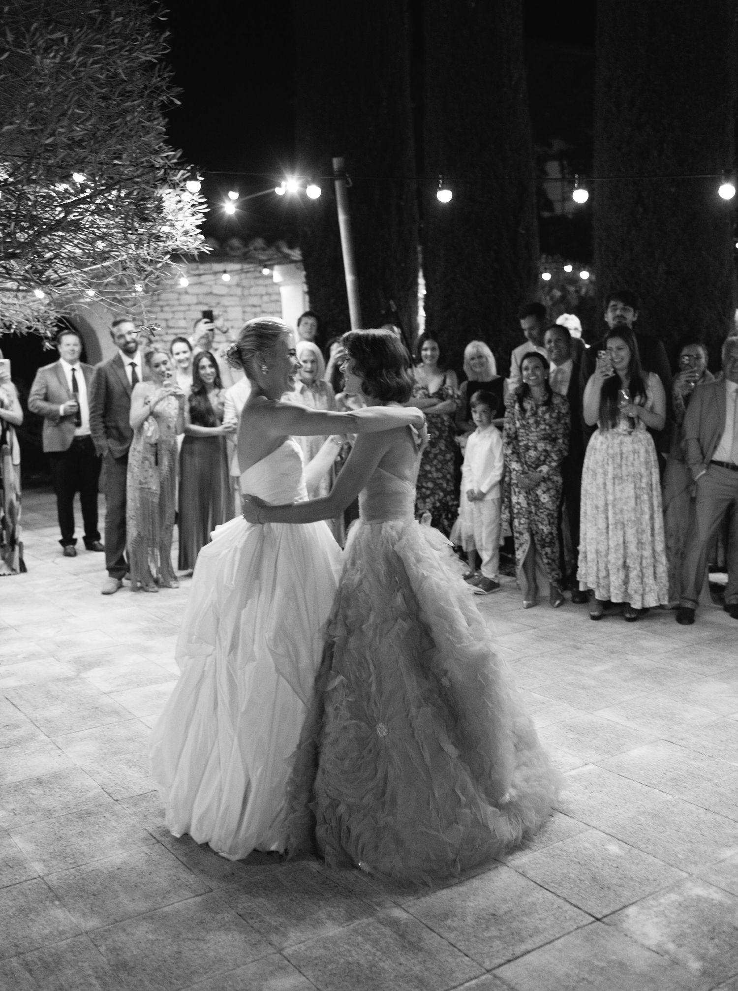 A group of wedding guests in formal attire dancing together on a stone patio during an evening reception celebration.