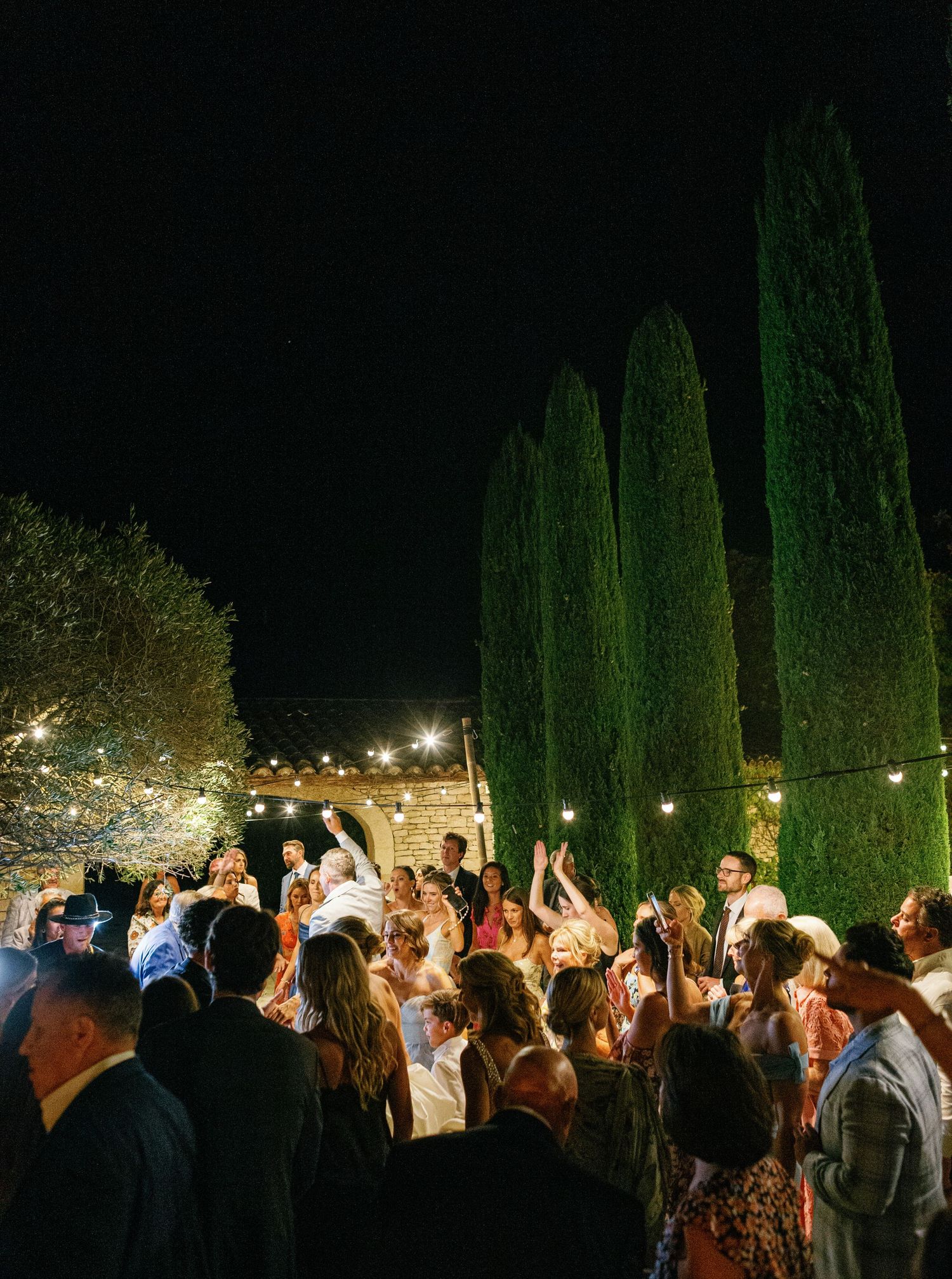 A crowd of people celebrating at a nighttime outdoor event with tall cypress trees and string lights in the background.