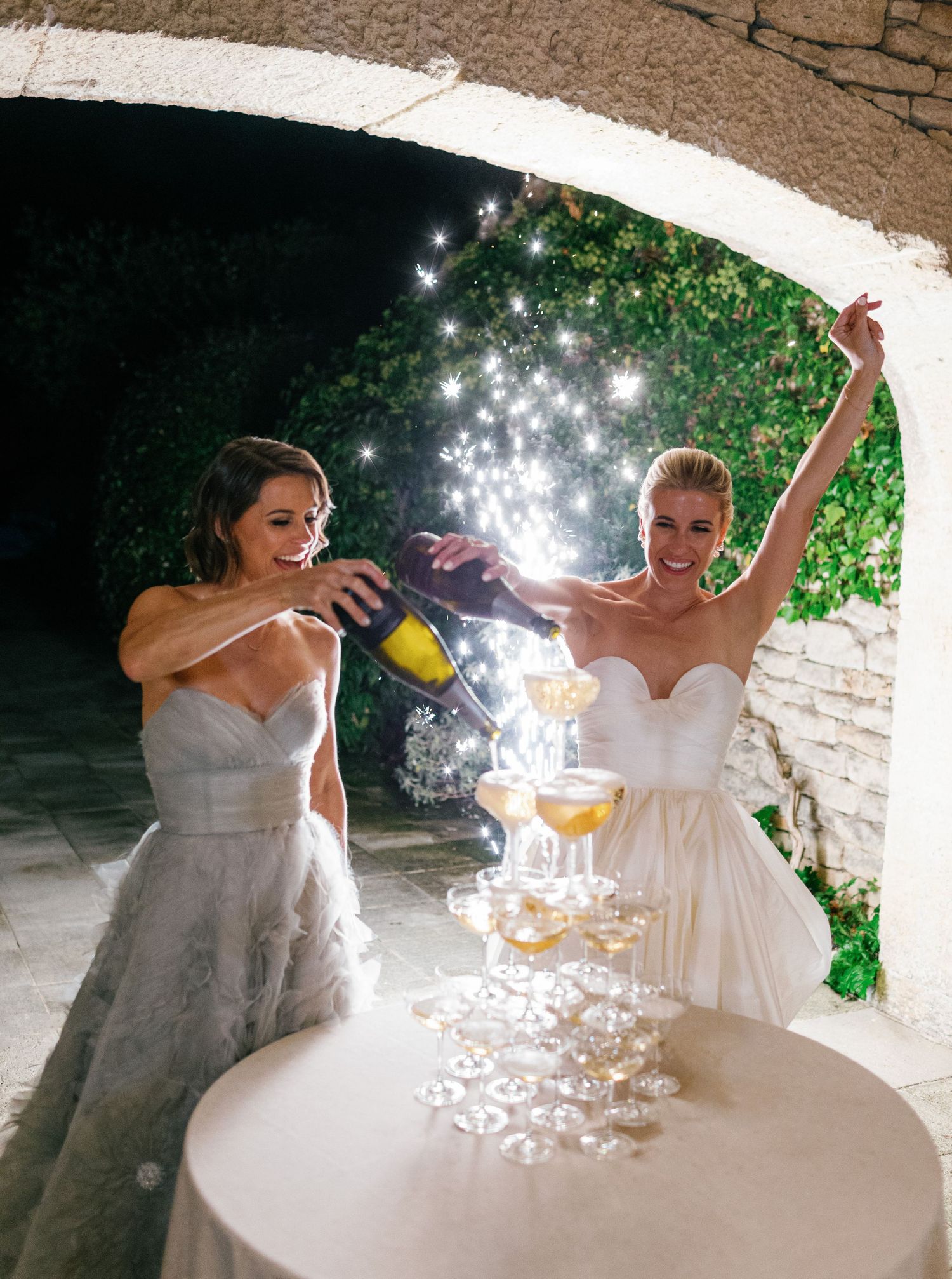 Wedding party members in white dresses celebrate with drinks and sparklers under a stone archway.