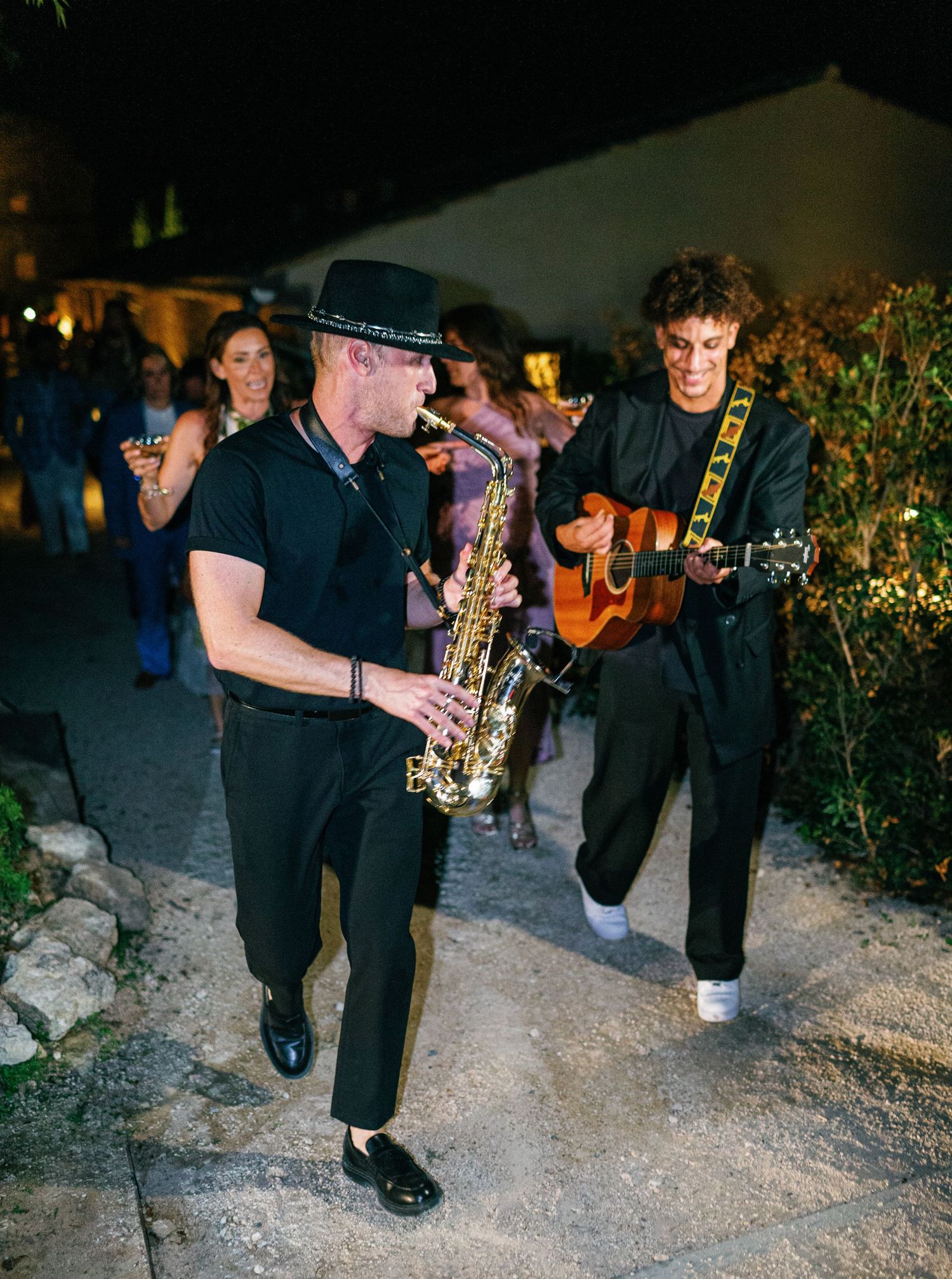 Musicians perform with saxophone and other instruments at an outdoor nighttime event on gravel ground.