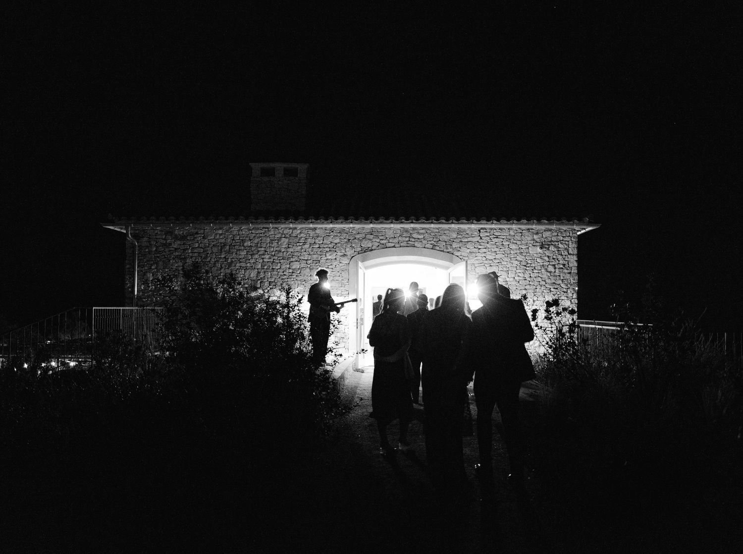 A silhouetted crowd stands against a dark night sky illuminated by bright spotlights at an outdoor event.