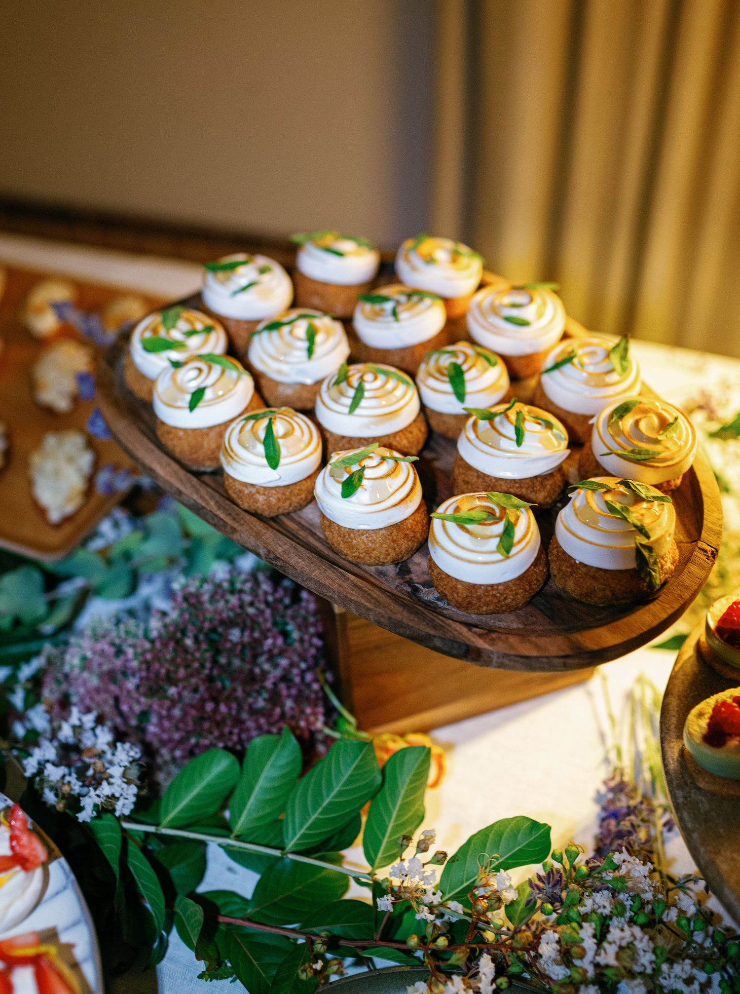 Cupcakes with white frosting and green garnish displayed on a rustic wooden platter beside fresh herbs and purple flowers.