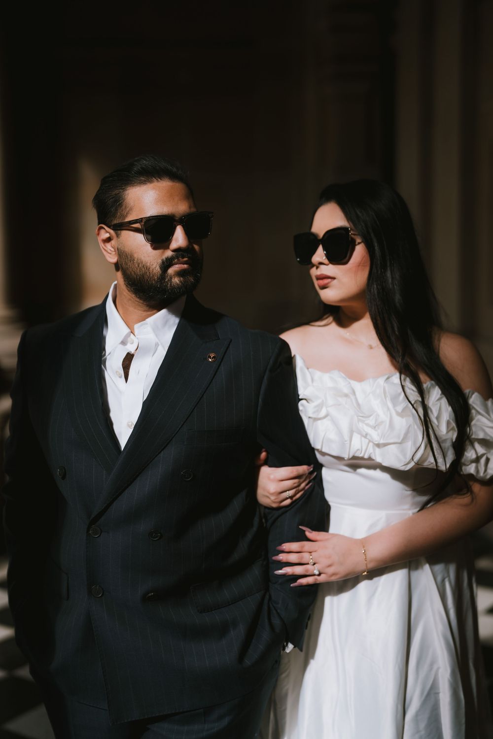Stylish couple in black and white attire pose together against dark background wearing sunglasses.