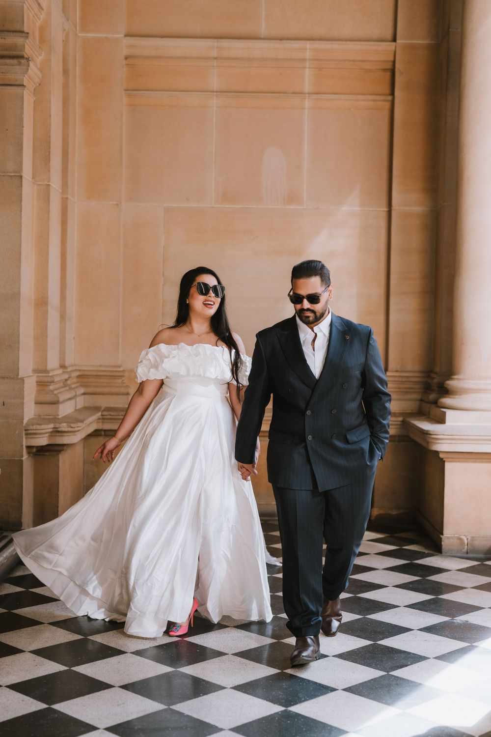 A couple in wedding attire walks together on a checkered floor in an elegant hallway with classical architecture.