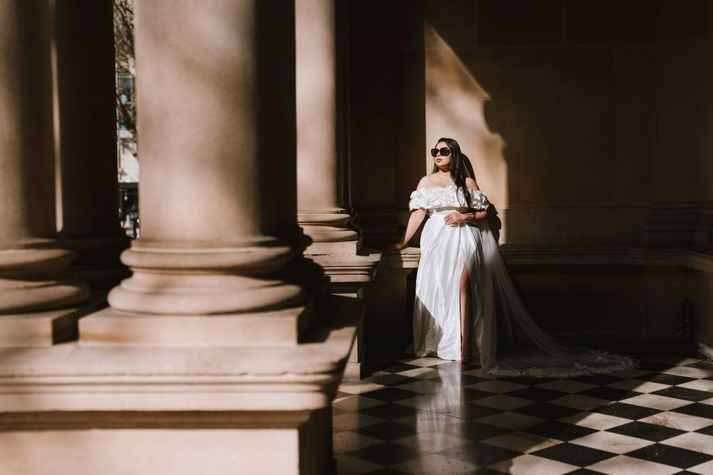 A bride in a white dress stands elegantly between classic stone columns in a dramatic black and white composition.