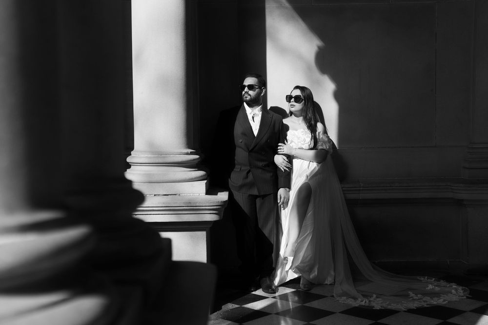 Elegant couple in formal wear standing between grand columns in moody lighting.