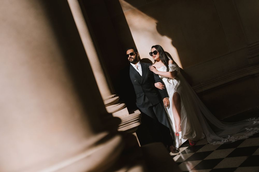 Elegant wedding couple in formal attire poses in dramatic lighting against a dark interior background.