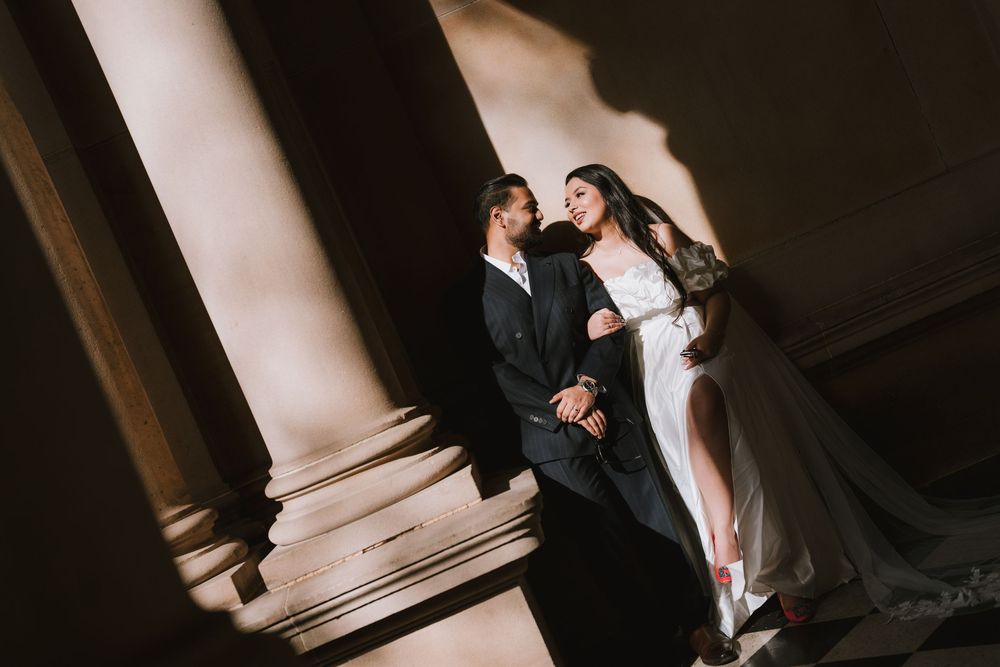 A couple in formal wedding attire poses together in a dramatic architectural setting with columns and shadows.