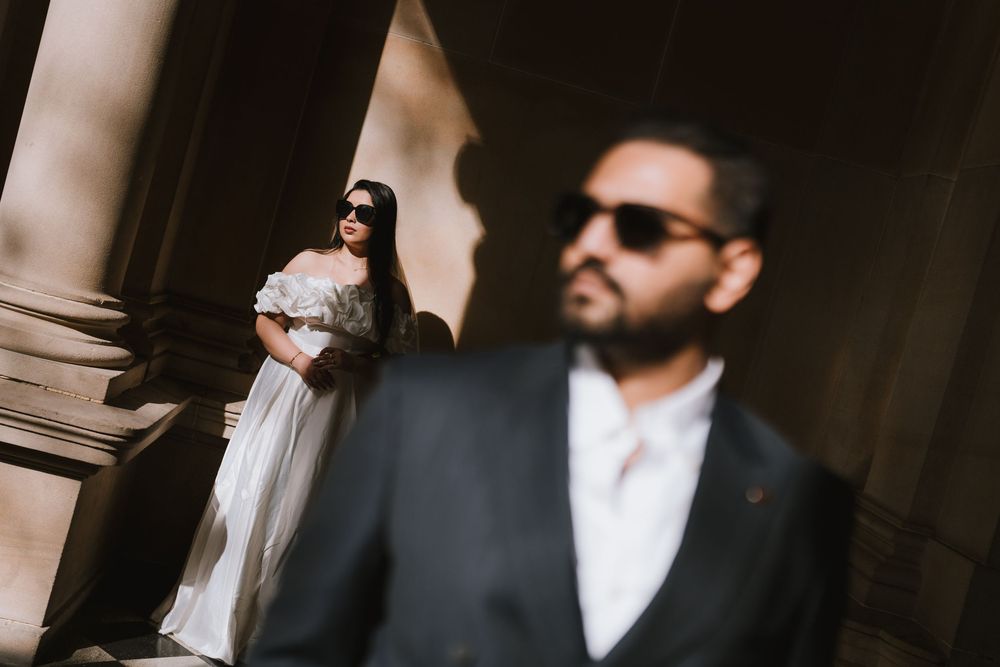 A couple in formal attire poses together against a dark background wearing sunglasses.