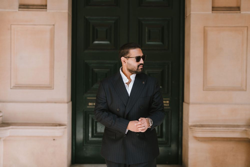 A person in a dark suit and sunglasses walks confidently past cream-colored columns in an elegant hallway.