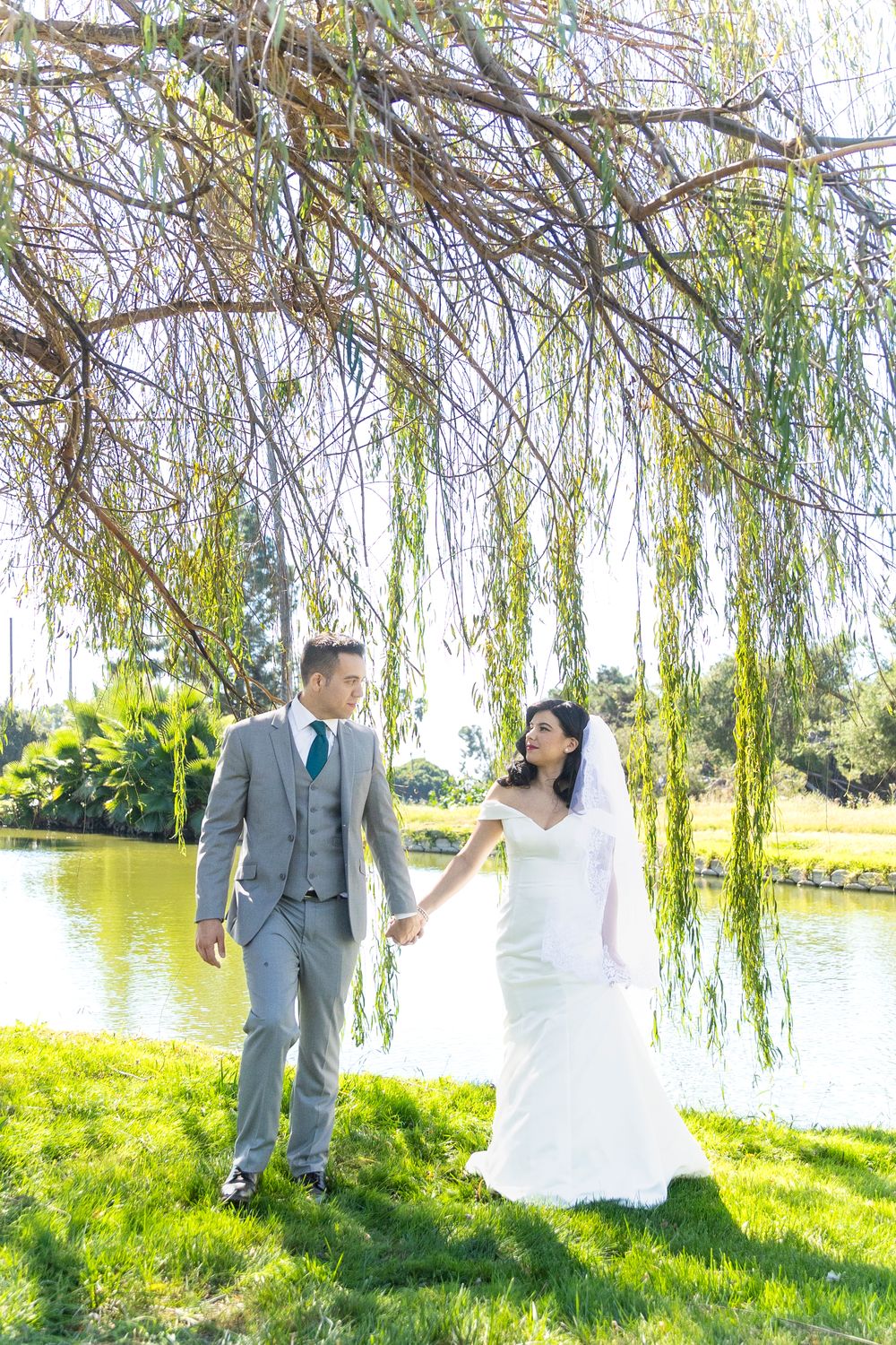A bride and groom walking while holding hands by a willow tree on their wedding day.