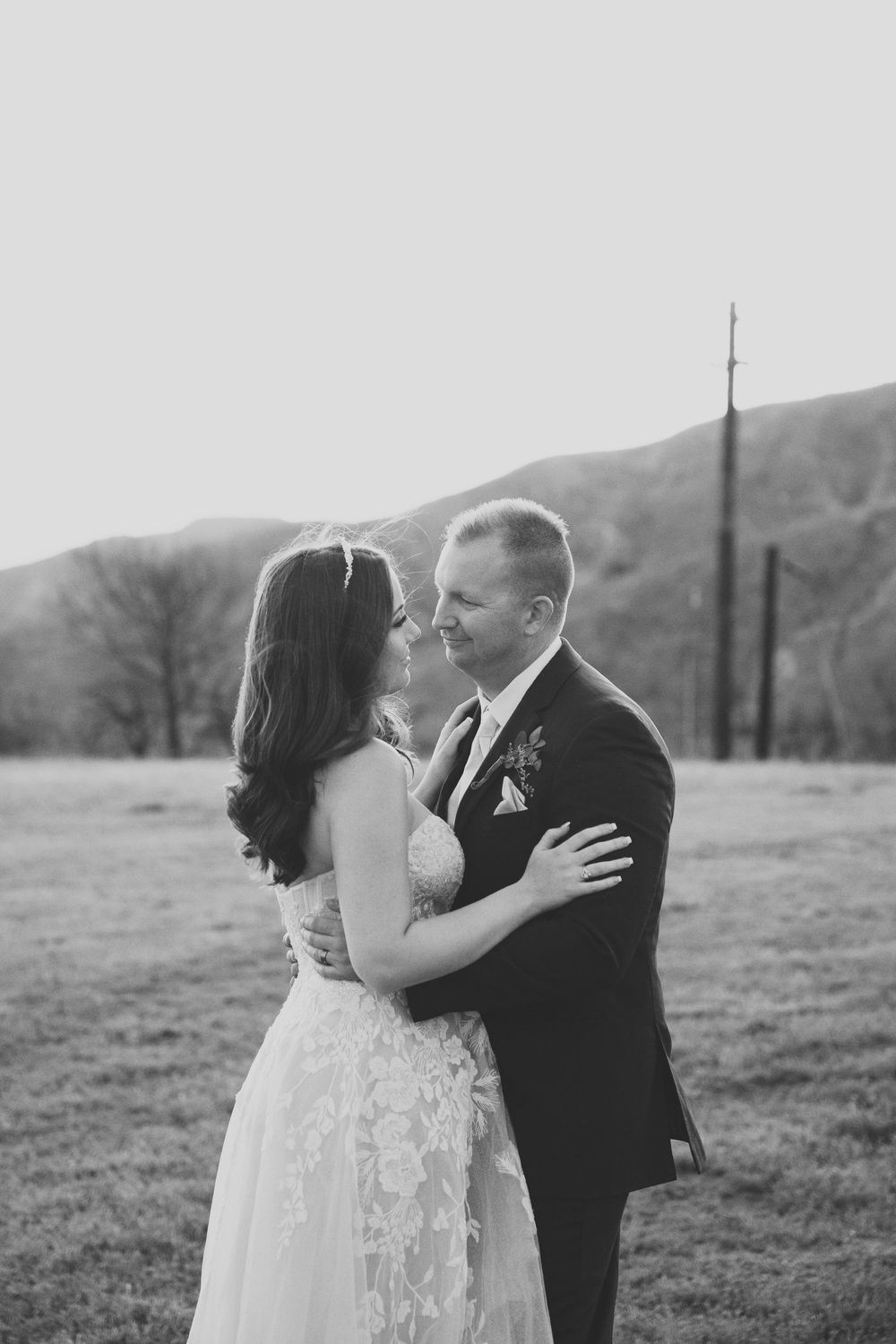 A black and white photo of a bride and groom on their wedding day holding one another in an open field by the mountains.