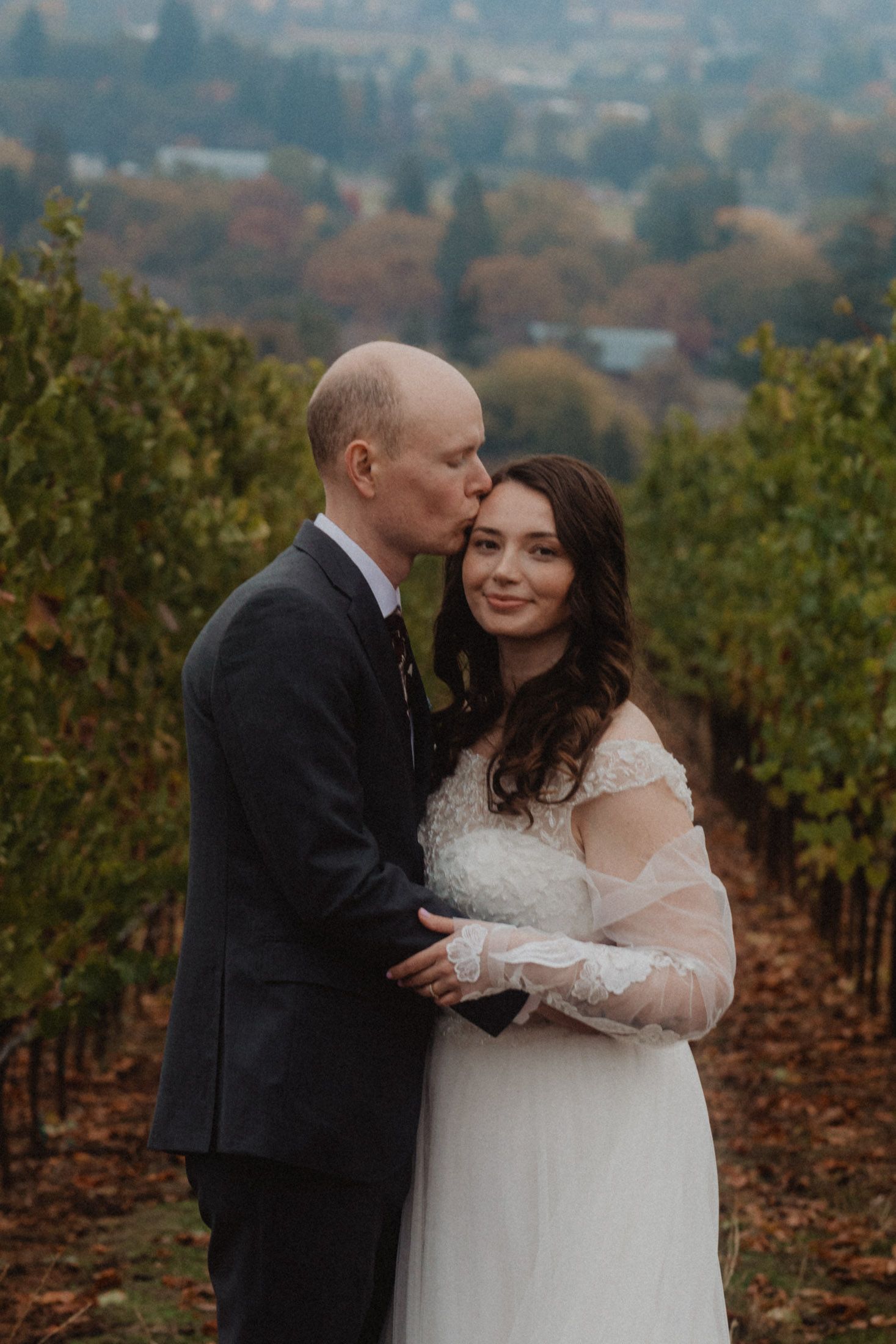 Romantic wedding couple embraces in autumn vineyard with rolling hills and misty landscape in background.