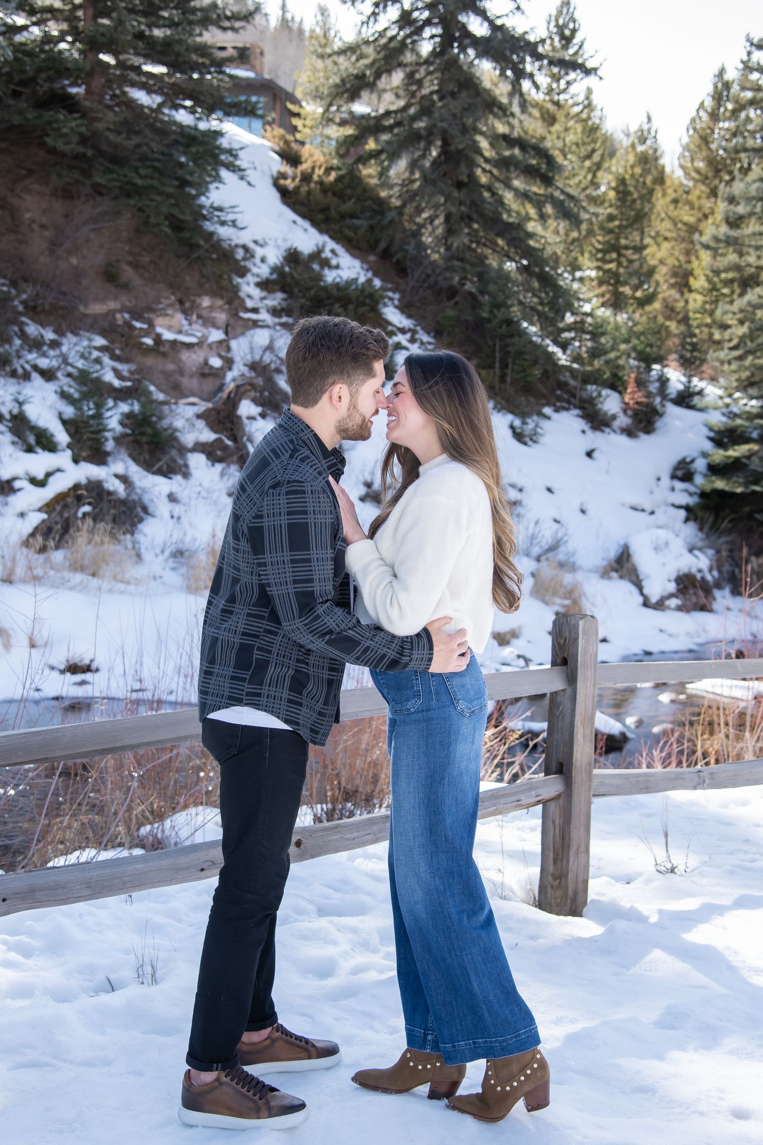 A  stylish couple embraces in a snowy winter forest setting with trees and a wooden fence in the background for Vail engagement photos