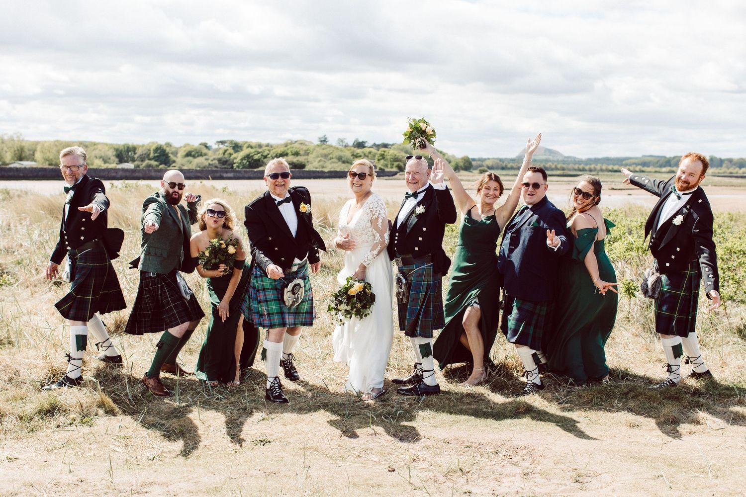 Wedding party members jump in unison on a grassy hillside while posing for a fun group photo.
