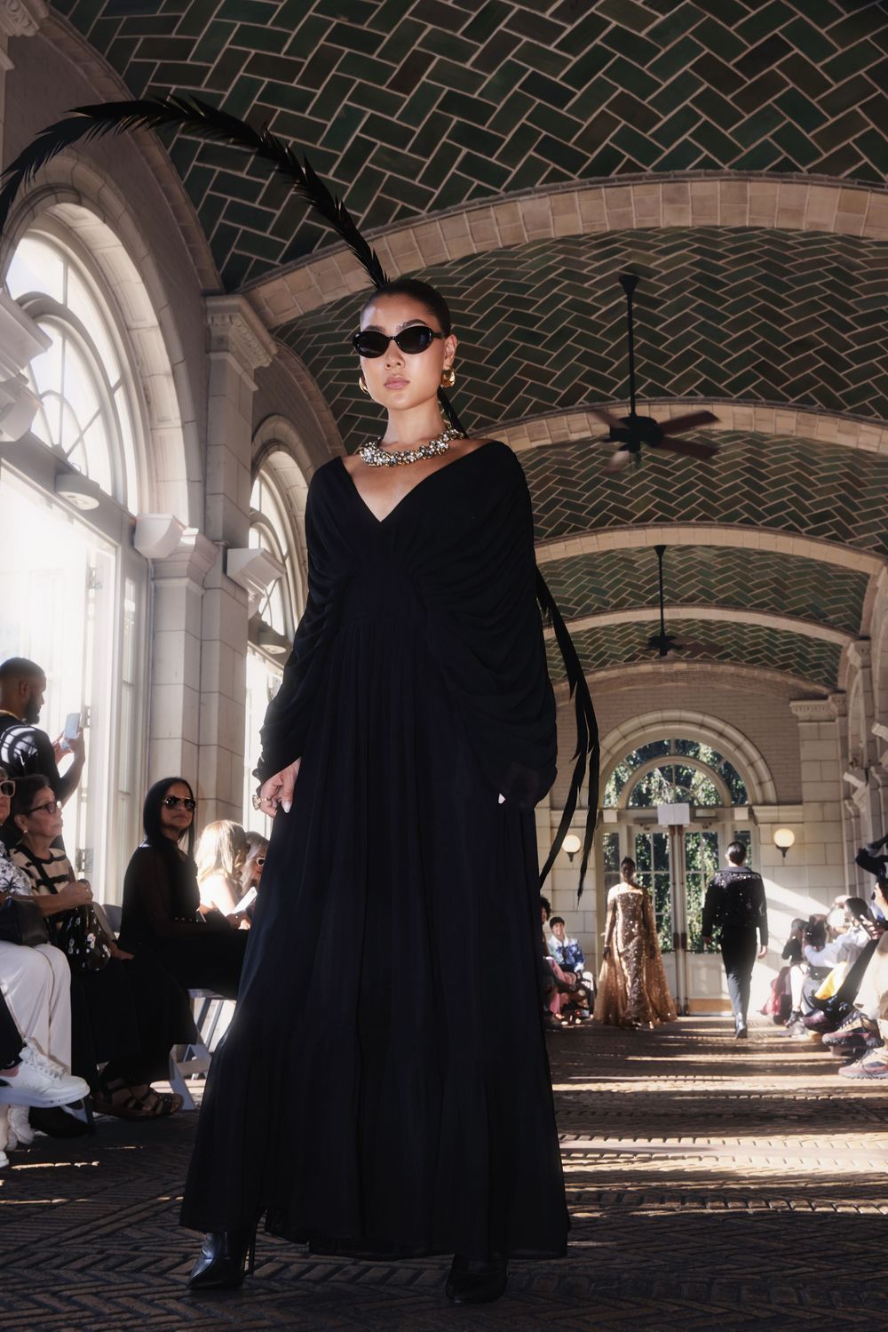 Models walk down a curved runway beneath an arched ceiling with geometric tile patterns during a fashion show.