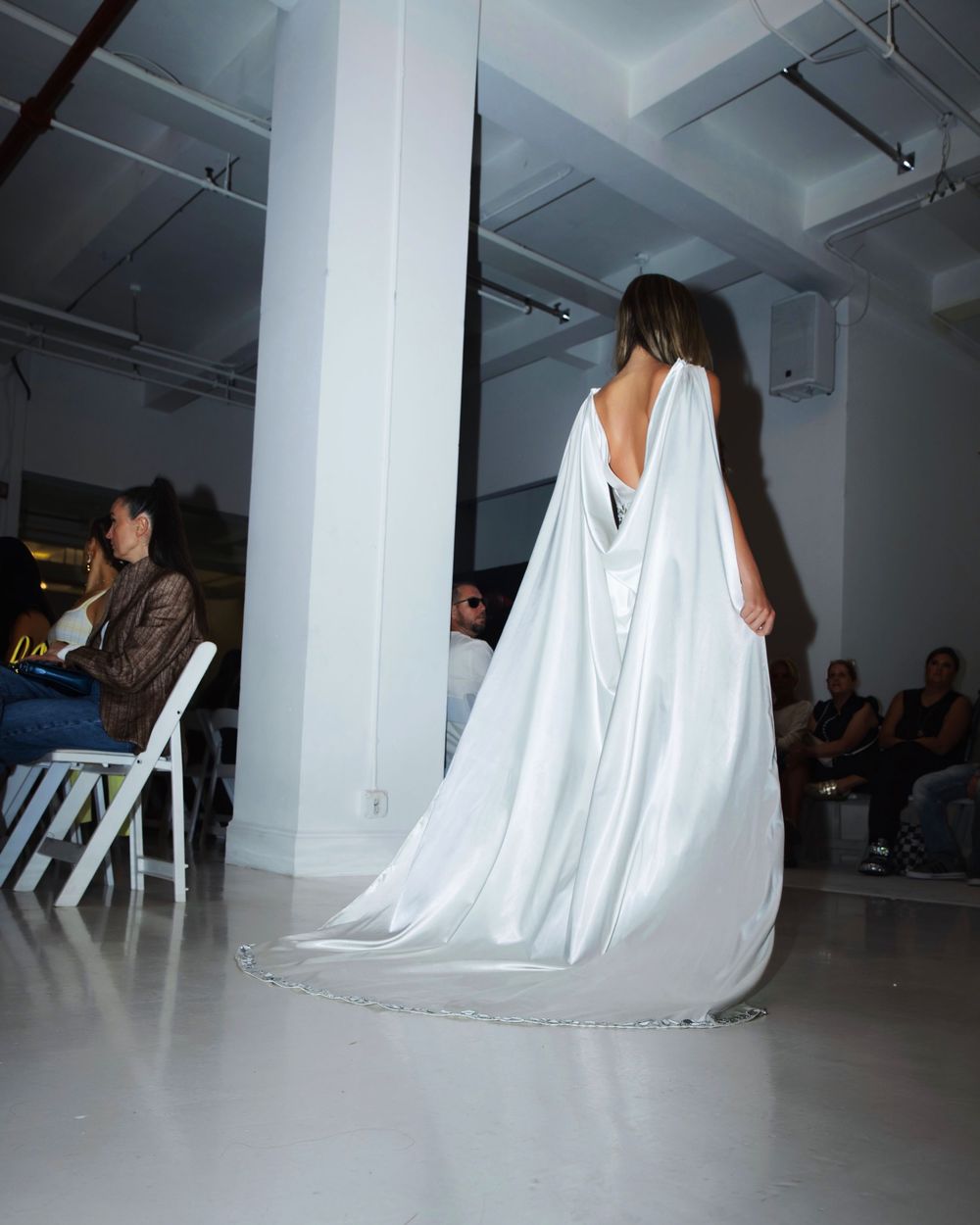A white sheet-covered figure sits on a chair in a minimalist gallery space with white walls and exposed ceiling.