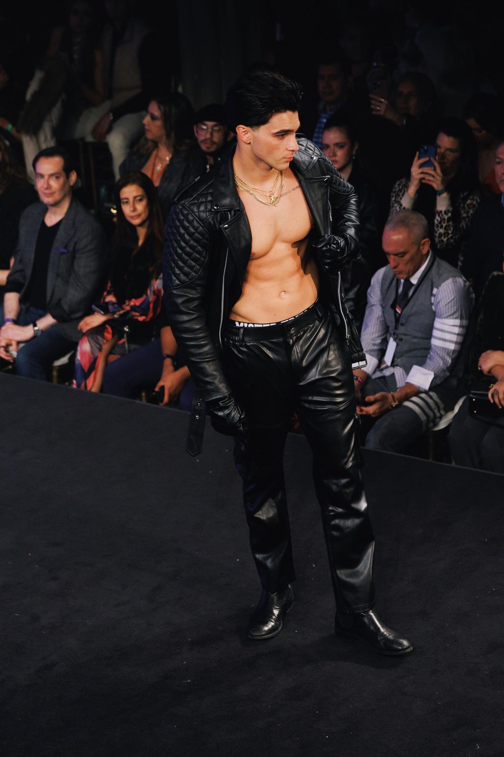Fashion model walks runway in black leather outfit during dark fashion show.