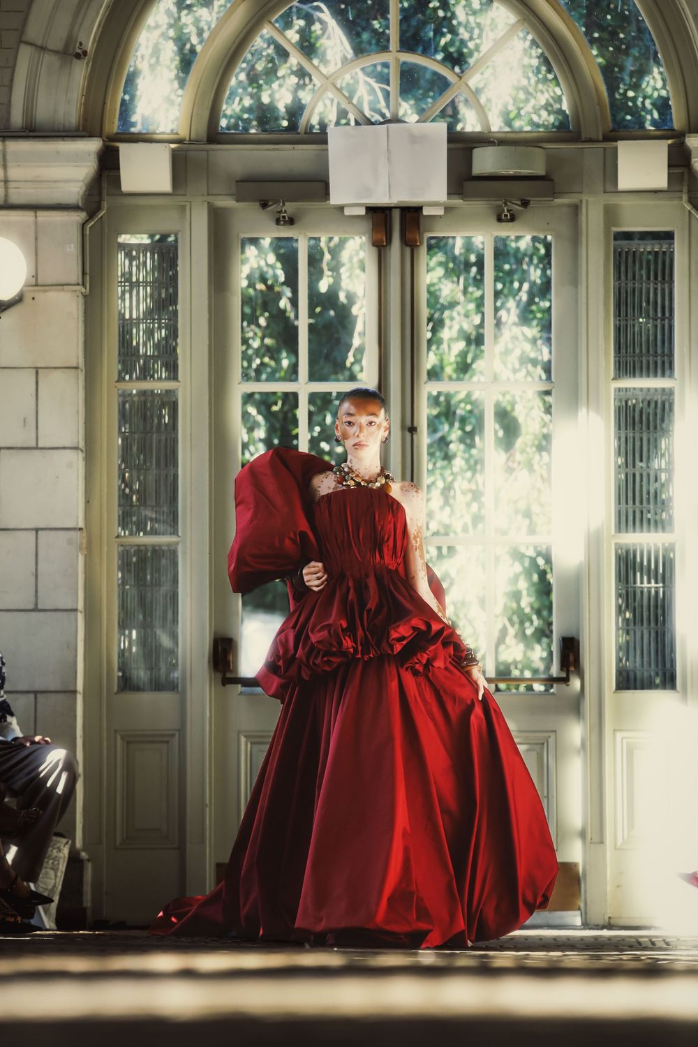 A figure in a flowing red Victorian-style dress moves gracefully through a sunlit columned hallway.