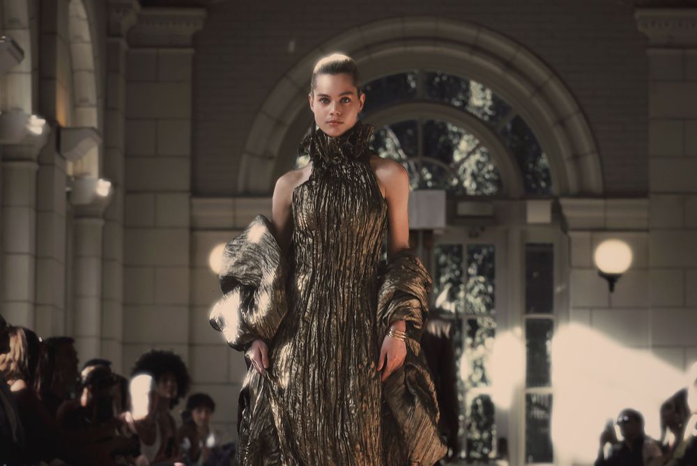 A person wearing a dramatic black and gold sequined gown walks down an ornate hallway during a fashion show.