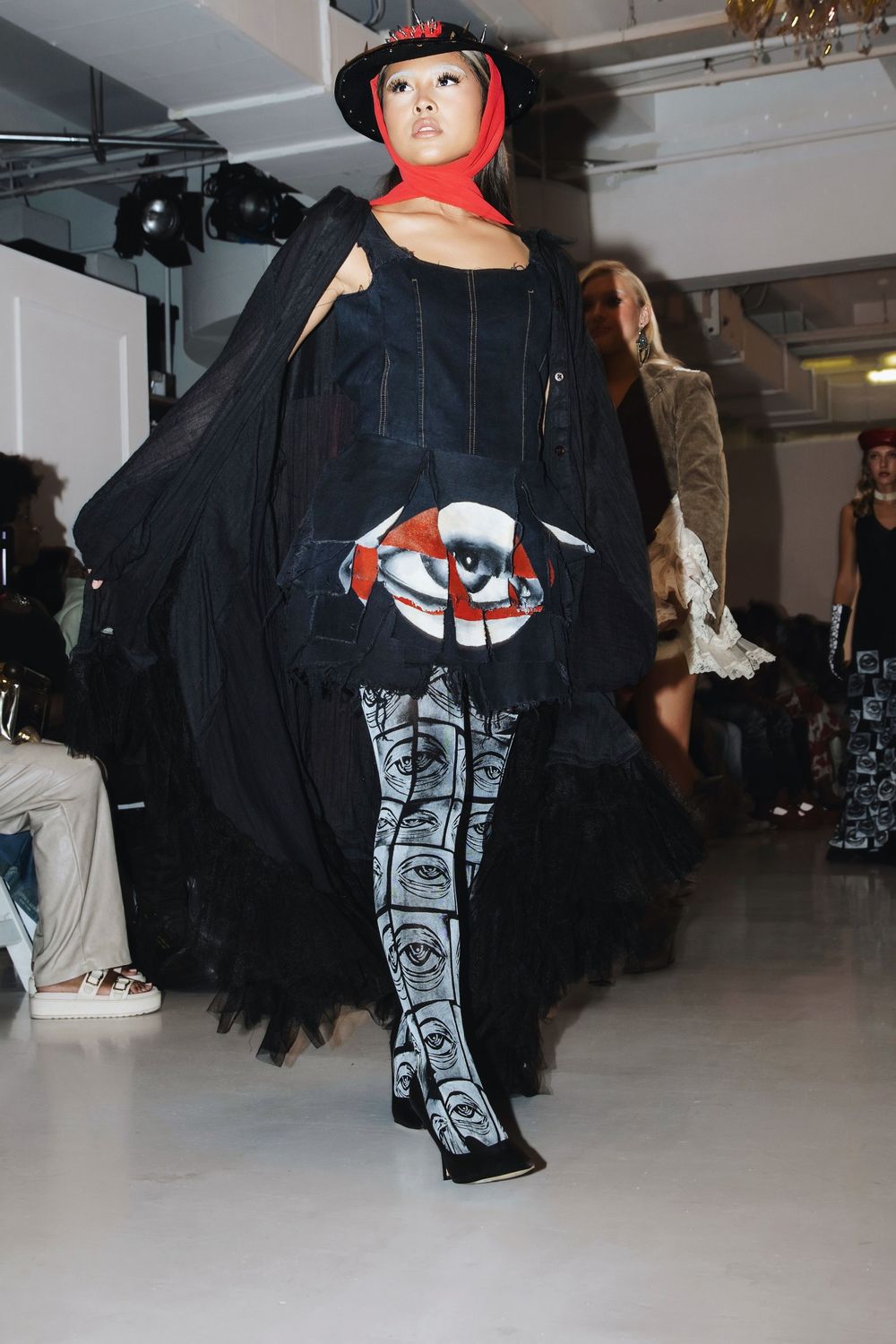 Fashion model wearing black cape with graphic print and red cap walks runway in industrial space.