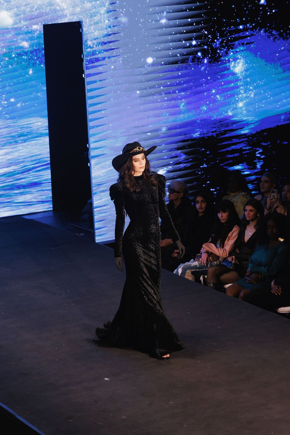 A model walks the runway in a dramatic black gown against a blue illuminated backdrop during a fashion show.