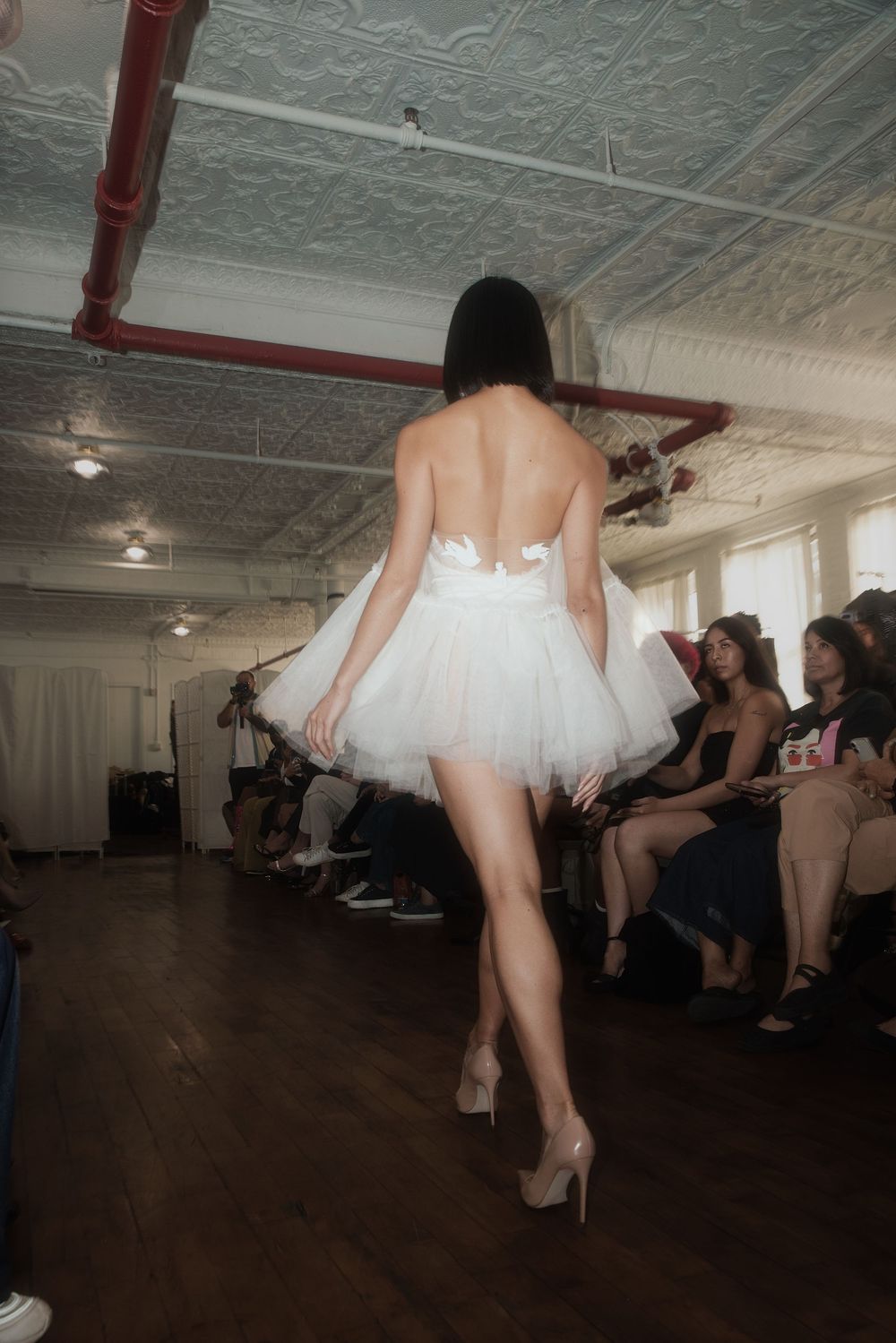 Model walks runway in short white tulle dress and heels at fashion show with exposed industrial ceiling.