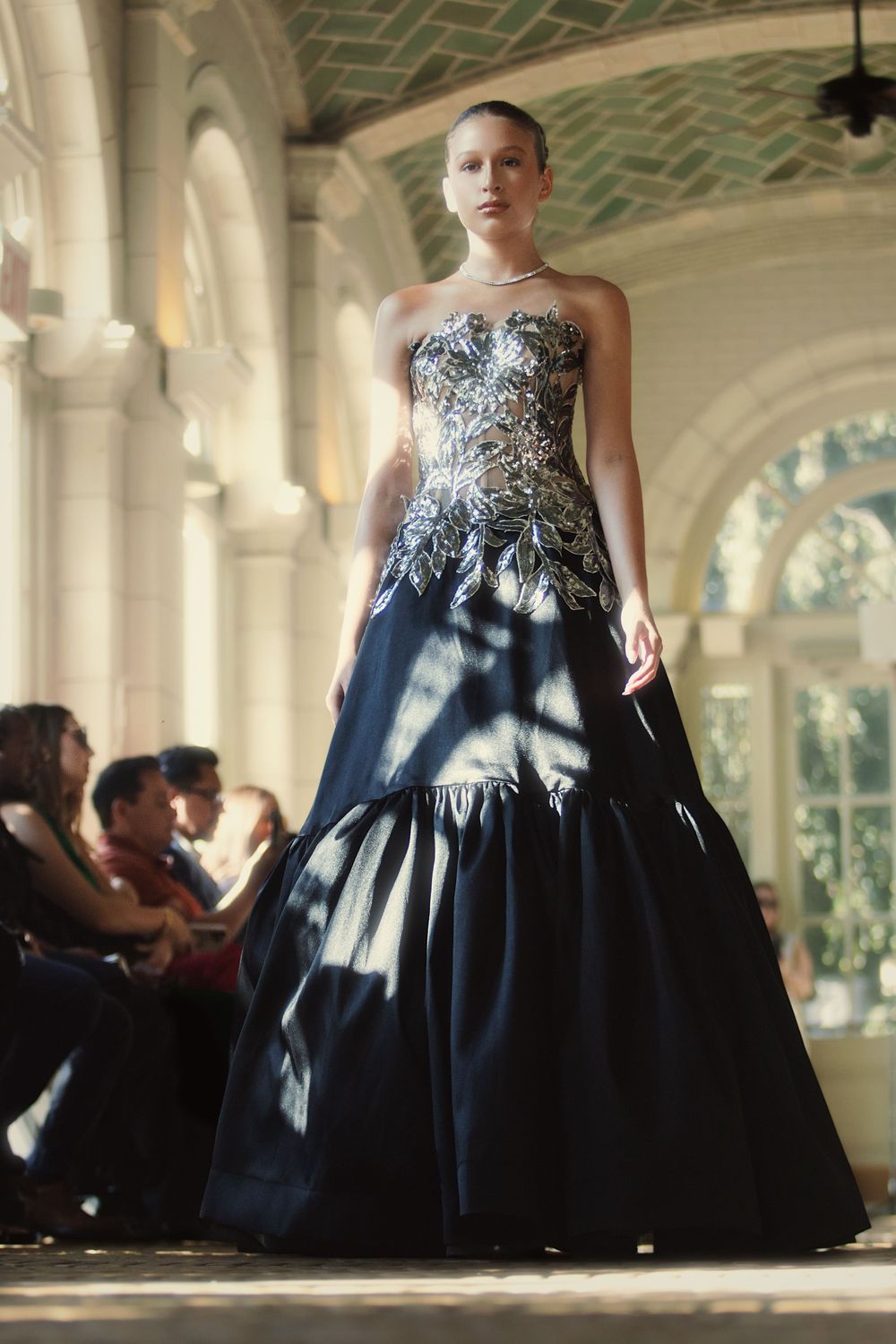 A model walks the runway in an elegant strapless evening gown with embellished bodice and navy blue full skirt in an ornate venue.