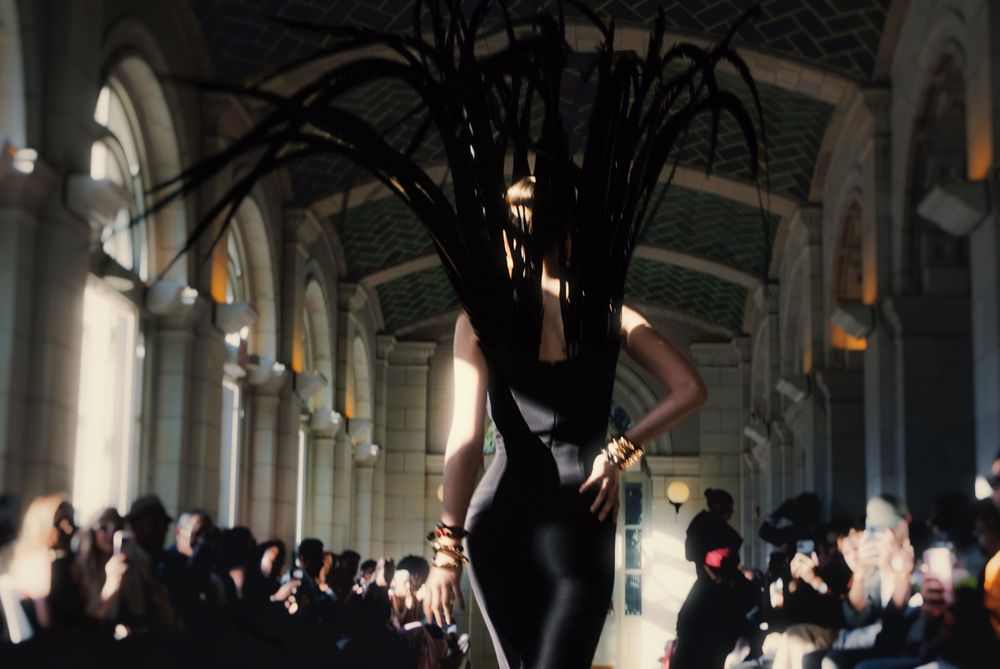 Fashion models walk down a runway in a grand historic building with ornate columns and arched architecture.