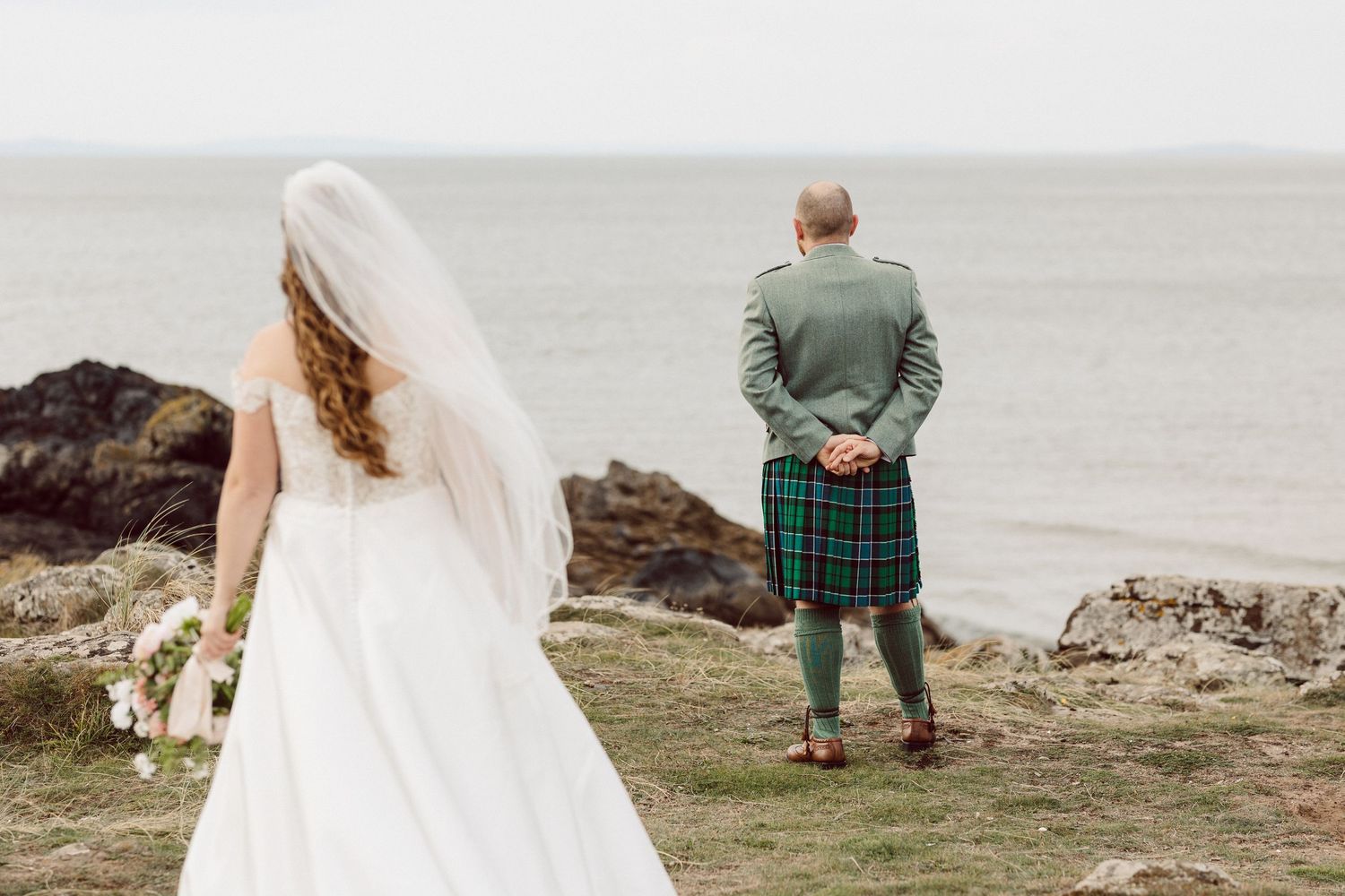 Couple stands at rocky ocean shore during cloudy day while wearing wedding attire.