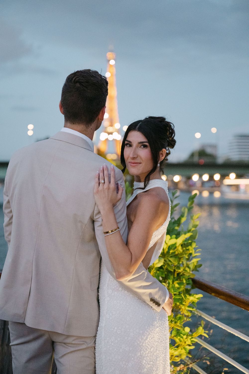 Newlyweds share an intimate moment at dusk on a waterfront deck with twinkling lights.