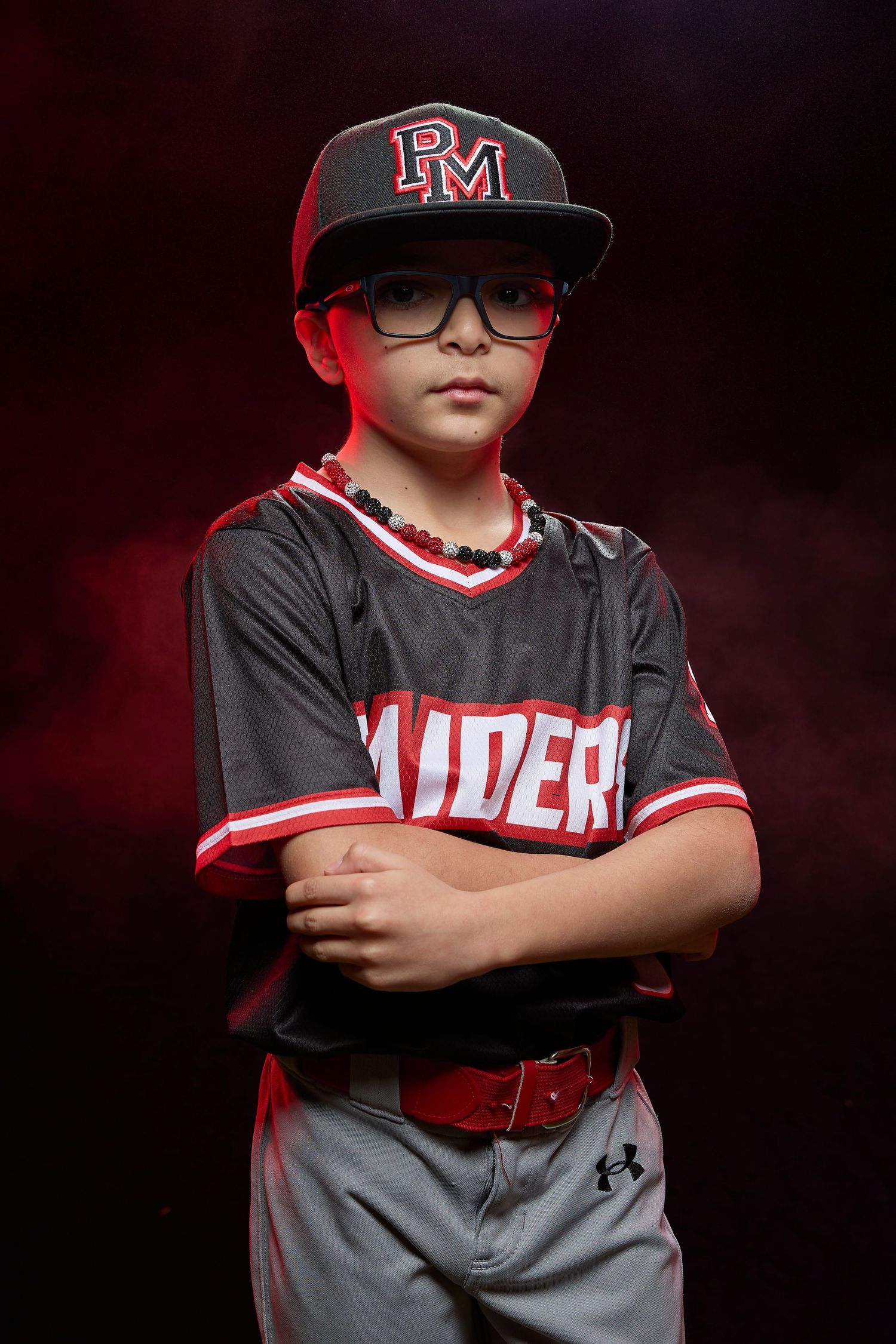 Youth baseball player in black and red uniform poses with arms crossed for dramatic sports portrait.