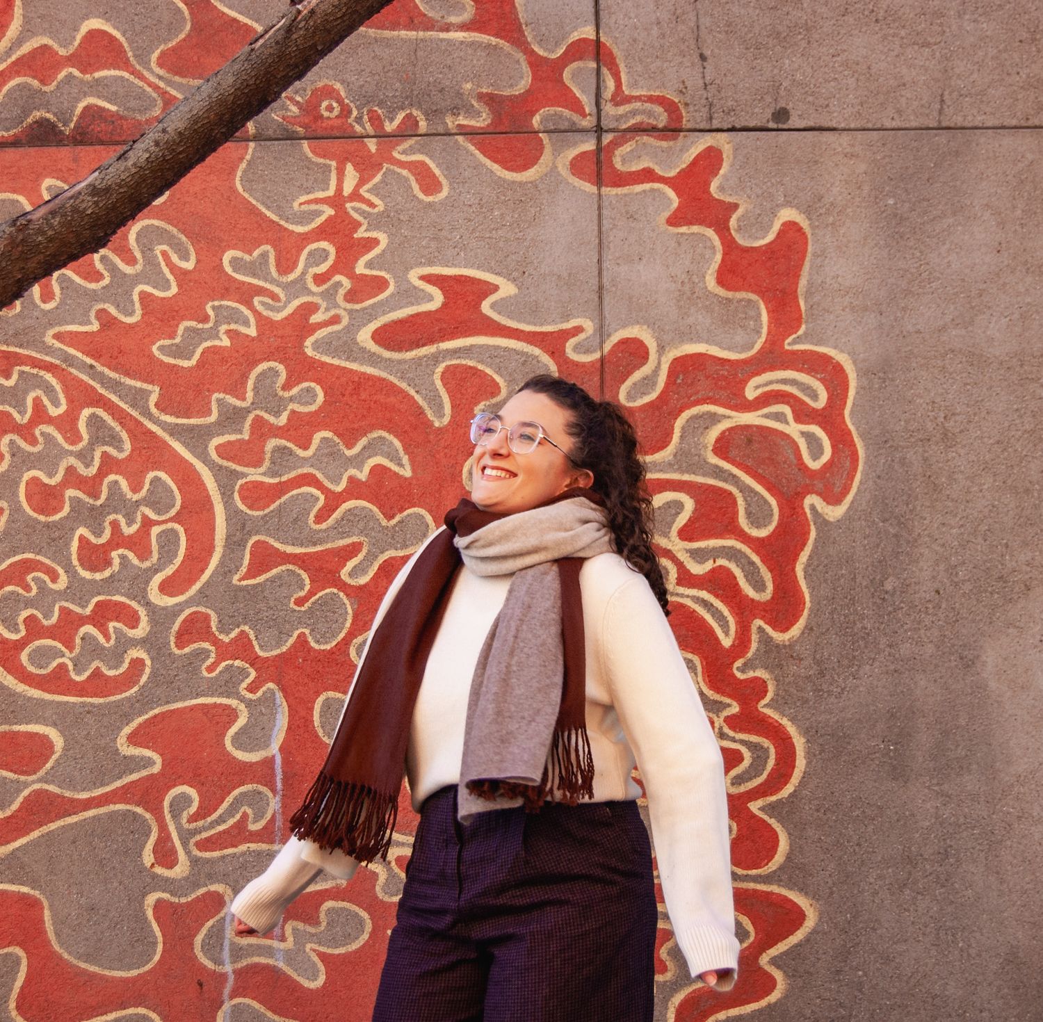 Smiling woman in cream sweater and scarf poses against vibrant red floral mural wall outdoors.