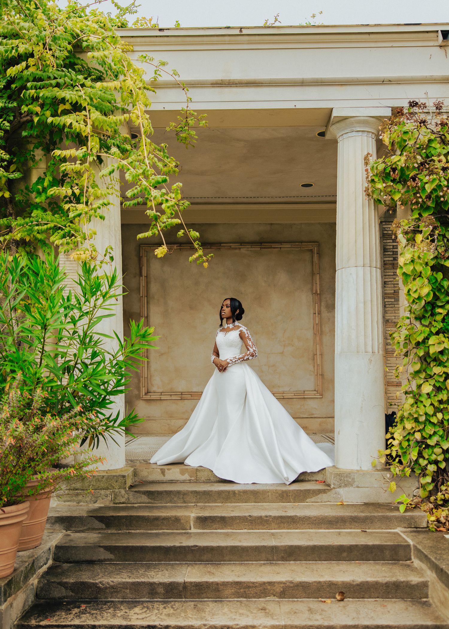 A bride in a flowing white gown poses elegantly on stone steps surrounded by lush greenery and white columns.