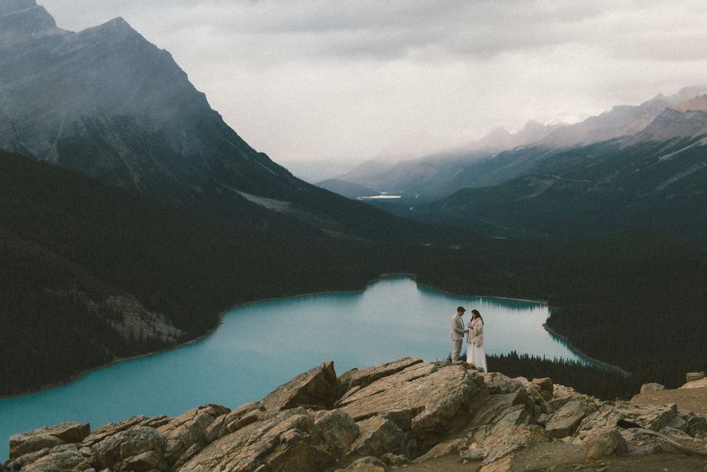 bride and groom hold each other on a rocky outcrop above a glacier blue lake with mountain peaks all around them at Peyto Lake in Banff