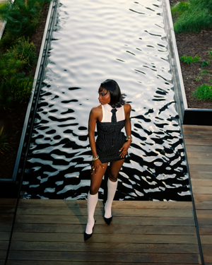 A person in a black and white outfit poses on a wooden deck against a textured metallic wall with wavy patterns.
