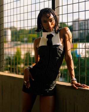 A person in black clothing poses against a metal grid fence with warm sunlight creating dramatic shadows.